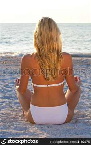 A beautiful young blond woman in a white bikini sits crosslegged on a beach at sunset