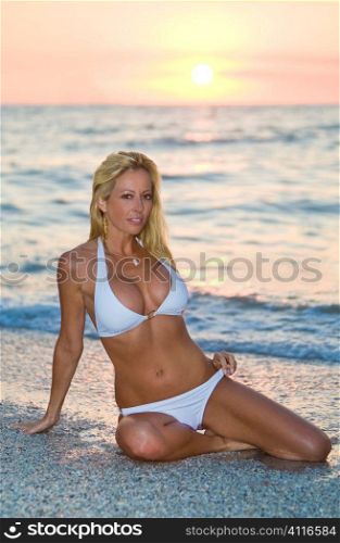 A beautiful young blond woman in a white bikini sits on a beach at sunset
