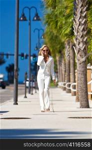 A beautiful young blond woman on the phone walking down a street lined with palm trees.