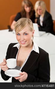 A beautiful young blond woman or businesswoman in a suit drinking coffee or tea in an office with her two female colleagues working on a laptop computer out of focus behind her