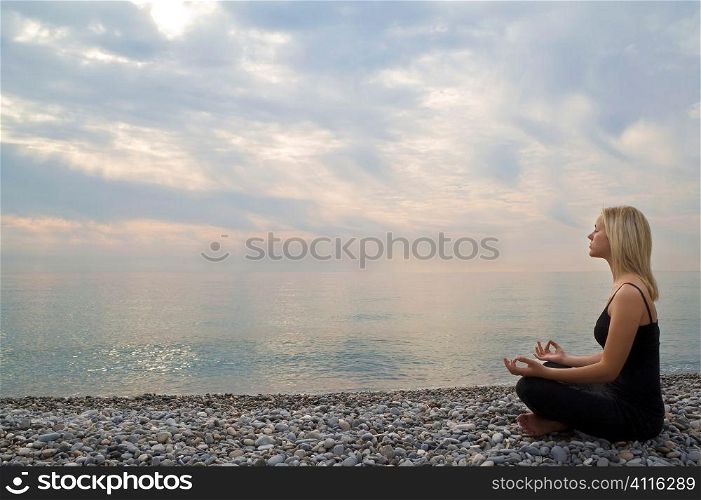 A beautiful young blond woman sitting cross legged in a yoga position on a beach at sunrise