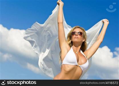 A beautiful young blond woman wearing a white bikini and sunglasses with white material blowing in the wind shot against a blue summer sky