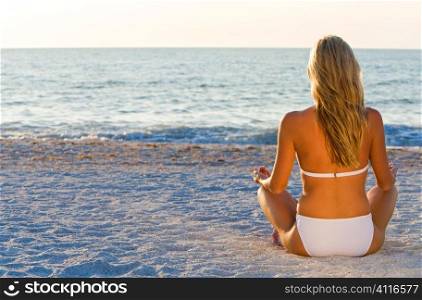 A beautiful young blond woman wearing a white bikini sitting cross legged on a beach at sundown