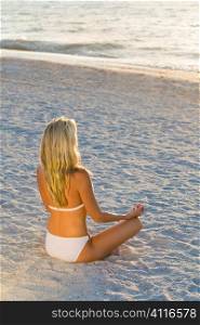 A beautiful young blond woman wearing a white bikini sitting cross legged on a beach at sundown