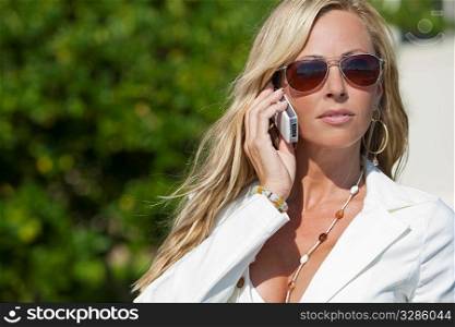A beautiful young blond woman wearing aviator sunglasses and a white suit talking on her cell phone in a sunny location