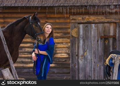 A beautiful young girl holds a horse by the bridle, against the background of a log wall