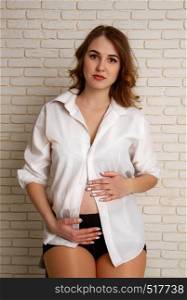 A beautiful young girl in her sixth month of pregnancy in a shirt of her husband and black dignified golf sits on a rough high stool against a brick wall. young pregnant girl