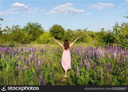 A beautiful young girl stands with her back in a field with purple lupins in a long dress and raises her hands up. A beautiful young girl stands with her back in a field with purple lupins in a long dress and raises her hands up.