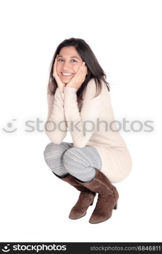 A beautiful young Hispanic woman in a knitted dress and brown bootscrouching on floor, the face in her hands, isolated on white background.. Happy woman crouching on floor.