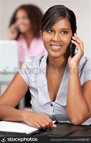 A beautiful young Indian Asian business woman with a wonderful smile in an office talking on her cell phone with her female colleague out of focus behind her.