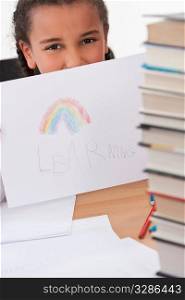 A beautiful young mixed race girl in a school classroom surrounded by books holds up a picture she has drawn of a rainbow with the word LEARNING underneath it.