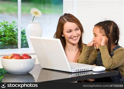 A beautiful young mother and her mixed race young daughter using a laptop at home in the kitchen.