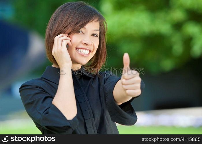 A beautiful young oriental woman with a wonderful smile outside chatting on her cell phone and giving a thumbs up.