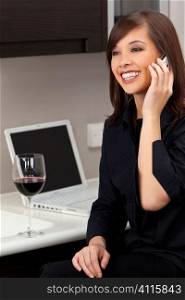 A beautiful young oriental woman with a wonderful toothy smile drinking red wine, chatting on her cell phone and using a laptop in her kitchen.