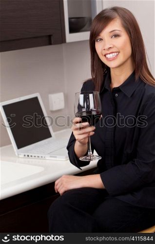 A beautiful young oriental woman with a wonderful toothy smile drinking red wine and using a laptop in her kitchen.