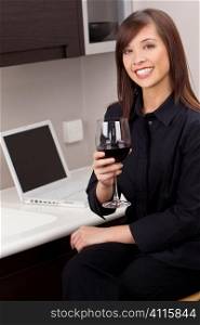 A beautiful young oriental woman with a wonderful toothy smile drinking red wine and using a laptop in her kitchen.