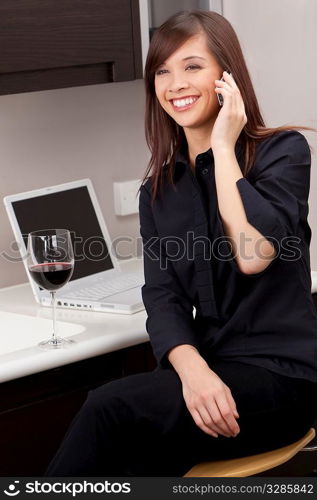 A beautiful young oriental woman with a wonderful toothy smile drinking redwine, chatting on her cell phone and using a laptop in her kitchen.