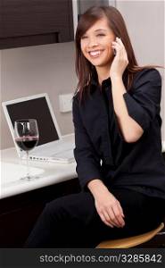 A beautiful young oriental woman with a wonderful toothy smile drinking redwine, chatting on her cell phone and using a laptop in her kitchen.