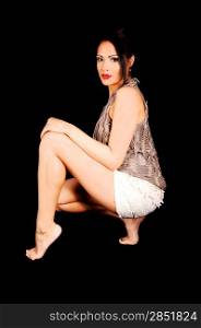 A beautiful young woman crouching in shorts and beige top for blackbackground, looking into the camera, barefoot.