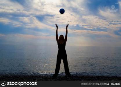 A beautiful young woman exercises with a ball on a beach with the early morning sun rising over a blue sea behind her.