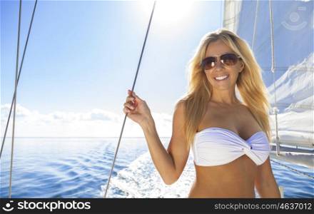 A beautiful young woman in a bikini and sunglasses standing a sail boat on a calm blue sea