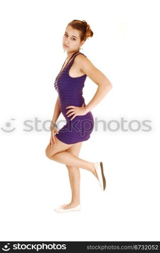 A beautiful young woman in a lilac dress, lifting up one leg,standing for white background with her hand on her hip.