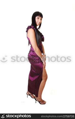 A beautiful young woman in a long burgundy evening dress standing inprofile in the studio, in heels and with long black hair, on white background.