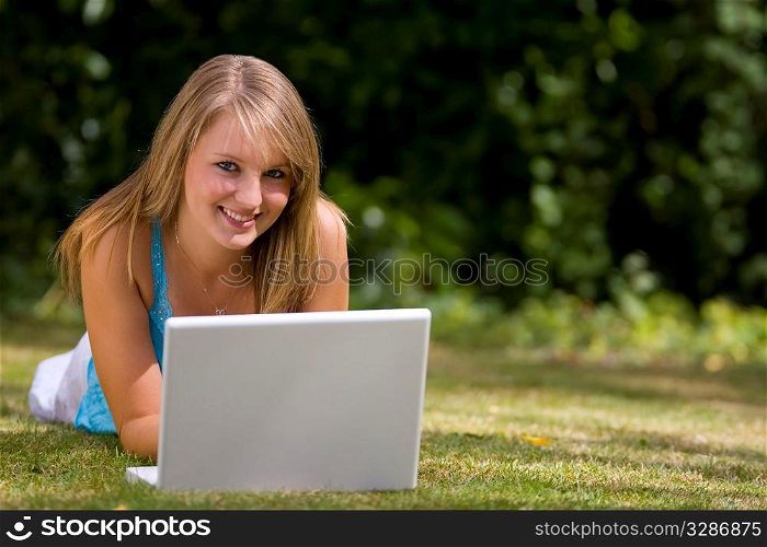 A beautiful young woman laying down in a grassy sunlit setting and working on her laptop