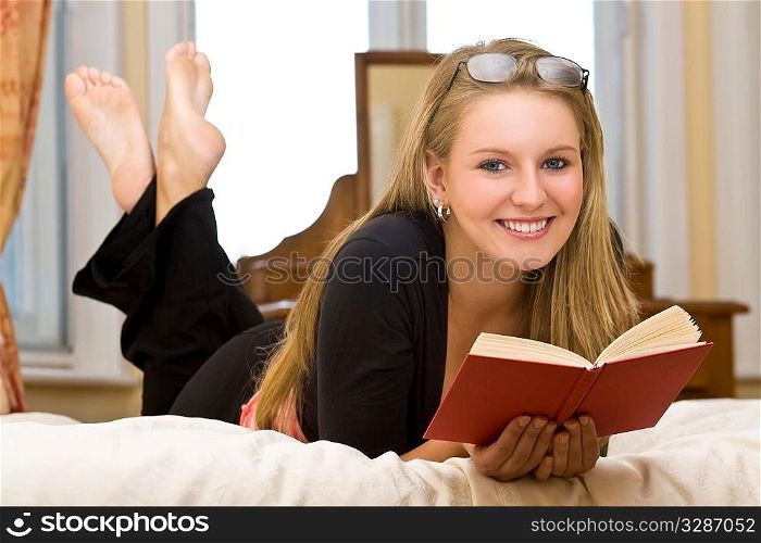 A beautiful young woman laying on her bed reading a book and smiling