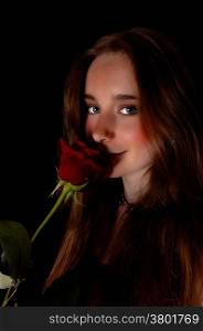 A beautiful young woman standing for black background holding a redrose to her face, looking into the camera.