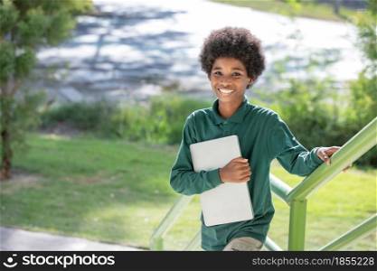 A black curly haired African boy hugs his laptop and smiles at the camera at the railing under the school building.Looking away.