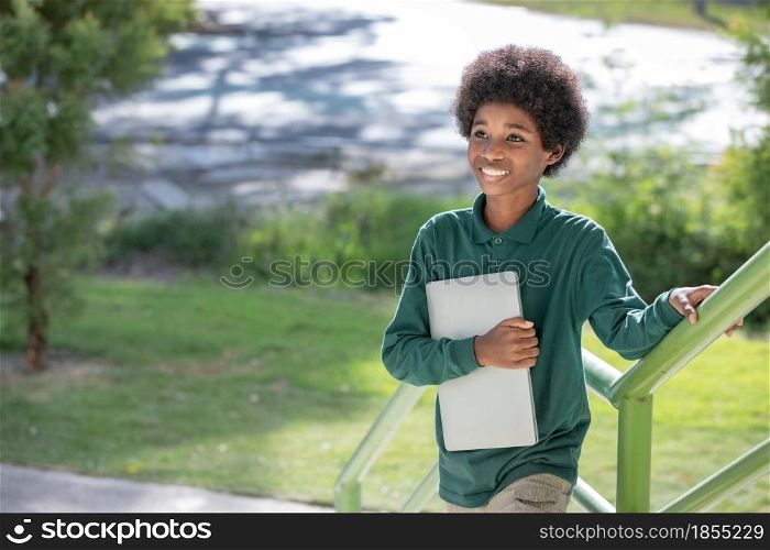 A black curly haired African boy hugs his laptop and smiles at the camera at the railing under the school building.Looking away.