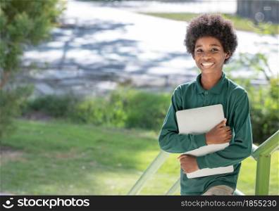A black curly haired African boy hugs his laptop and smiles at the camera at the railing under the school building.Looking at camera.