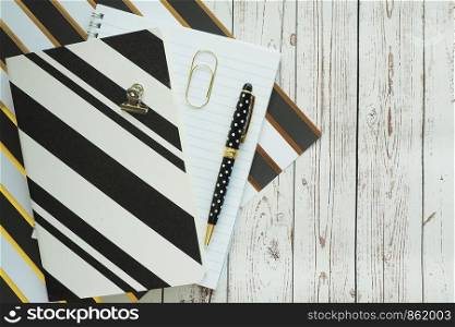 A black pen with polka dots, a notebook and a notebook, paper with black and gold stripes, paper clips, on a plain wooden background. Space for text.