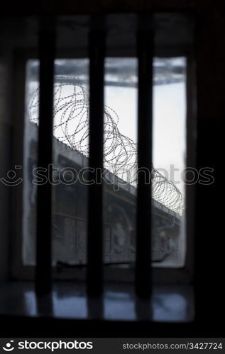 A bleak and dismal view through a prison window. The bars over the window with the only view of coils of barbed wire on a wall is symbolic of the lack of freedom for prisoners.