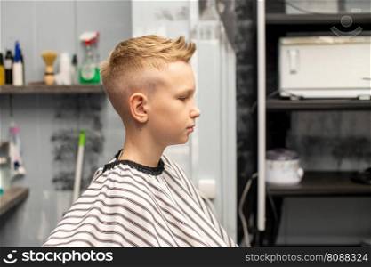 A blond boy is sitting in a barbershop, getting his hair done. A boy is sitting in a barbershop, getting his hair done