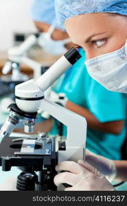 A blond female medical or scientific researcher or doctor using her microscope in a laboratory with her Asian colleague out of focus behind her.