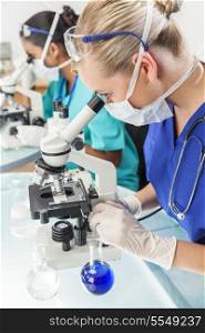 A blond female medical or scientific researcher or doctor using her microscope in a laboratory with her Asian colleague out of focus behind her