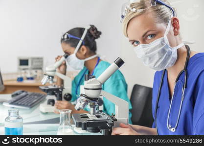 A blond female medical or scientific researcher or doctor using her microscope in a laboratory with her Asian colleague out of focus behind her.