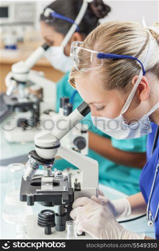 A blond female medical or scientific researcher or doctor using her microscope in a laboratory with her Asian colleague out of focus behind her.