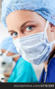 A blond female medical or scientific researcher or doctor wearing a surgical mask in a laboratory with her Asian colleague using her microscope out of focus behind her.