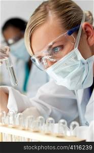 A blond medical or scientific researcher or doctor using looking at a clear solution in a laboratory with her Asian female colleague out of focus behind her.