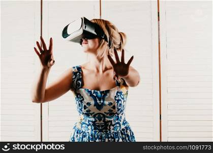 A blonde pretty woman wearing virtual reality glasses, pointing her hands in the air, light wooden background