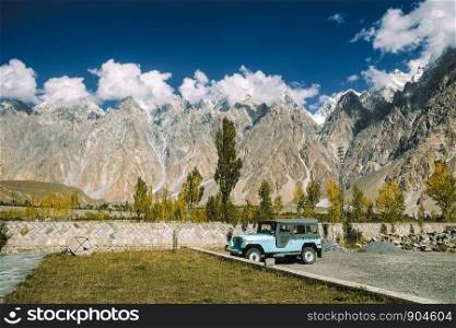 A blue car parking near Passu cones massif mountain peaks in Karakoram range with clouds in autumn season. Gojal Hunza, Gilgit Baltistan, Pakistan.