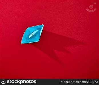 A blue origami paper boat presented on a red background with space for text. Top view. Handcraft paper blue ship isolated on red background with reflection of shadows and copy space. Top view