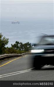 A blurred motor vehicle speeds by in the foreground on a mountain pass road with an oil drill platform in the ocean behind.