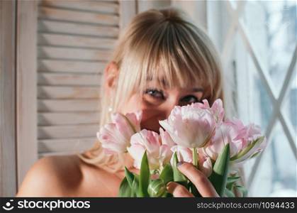A blurred view of a young blonde woman holding a bouquet of fresh pink flowers by the window, smiling, soft focused flowers