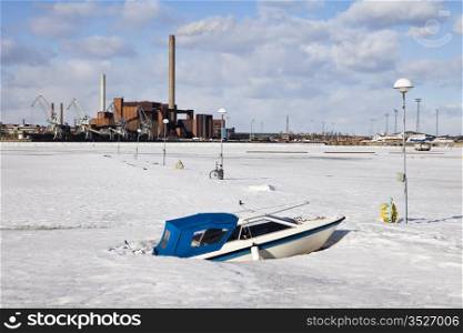 A boat is stranded in the frozen harbor of Helsinki in the middle of winter. In the background, an industrial plant is operating.
