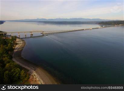 A boat moves along heading south going under the Hood Canal Bridge near the Olympic Mountain Range in the background