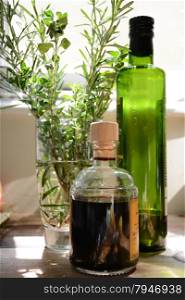 a bottle of olive oil and a bottle of balsamic vinegar next to a glass filled with rosemary and oregano